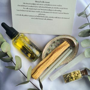 Puede incluir: Un kit de ritual de luna llena con una tarjeta, una botella de aceite, una concha, un palo santo, velas, una varita de selenita y un pequeño frasco de hierbas. La tarjeta tiene texto en holandés.