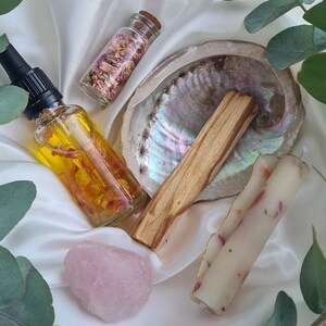 Op de afbeelding: Een assortiment spirituele en wellness-artikelen, waaronder een glazen fles olie met bloemblaadjes, een klein potje met gedroogde bloemblaadjes, een rozenkwartskristal, een houten palo santo-stok, een zeeschelp en twee kaarsen met bloemblaadjes.