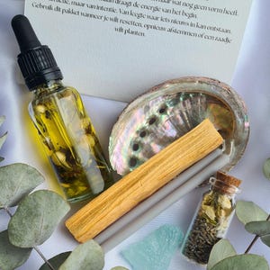 Puede incluir: Una colección de artículos espirituales que incluyen una botella de vidrio con cuentagotas, una concha, un palo santo, dos varillas de incienso grises, un pequeño frasco de vidrio con un corcho y un cristal azul. El texto en la tarjeta dice "Ritueel nieuwe maan".
