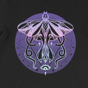 May include: Black t-shirt with a graphic of a moth with a pink and purple gradient, surrounded by a circle with a geometric pattern and a single eye in the center.