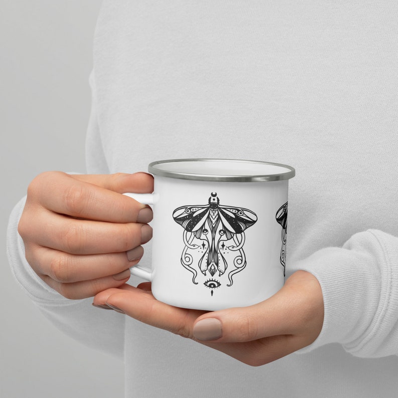 May include: White enamel mug with a black and white illustration of a moth with a crescent moon and an eye design.