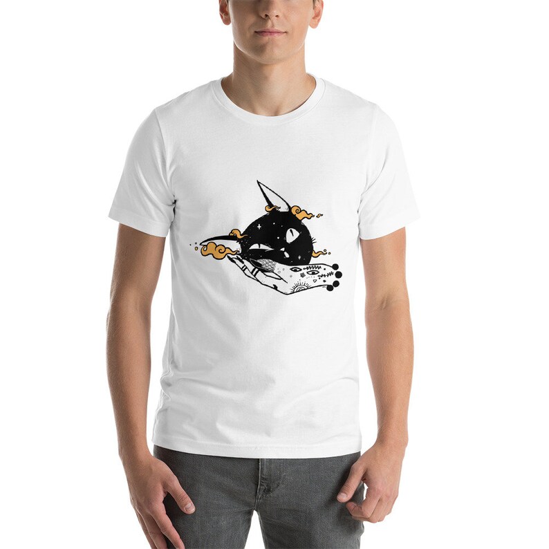 May include: White t-shirt featuring a black and white illustration of a hand holding a black cat's head with a crescent moon shape.