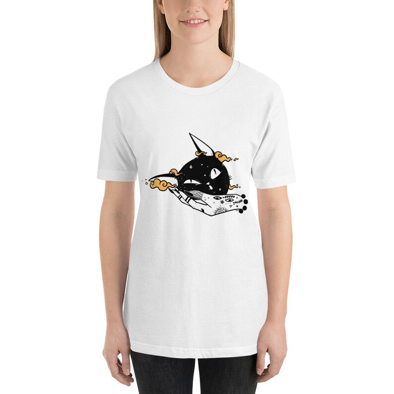 May include: White t-shirt featuring a black and white illustration of a cat's head with stars inside, held by a hand with a tattoo of eyes and other symbols.