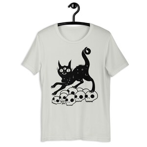 以下が含まれることがあります： 3つの目を持ち、星形の体をした黒い猫が、白い頭蓋骨の山の上に立っています。猫は口を開けて前を向いています。猫は黒で縁取られ、頭蓋骨は黒で塗りつぶされています。