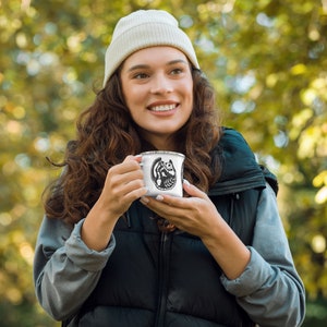 May include: A white enamel camping mug with a black and white illustration of a lemur on it. The mug is being held by a person wearing a white beanie and a black vest.