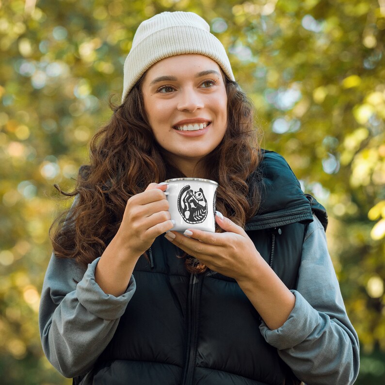 May include: A white enamel camping mug with a black and white illustration of a lemur on it. The mug is being held by a person wearing a white beanie and a black vest.