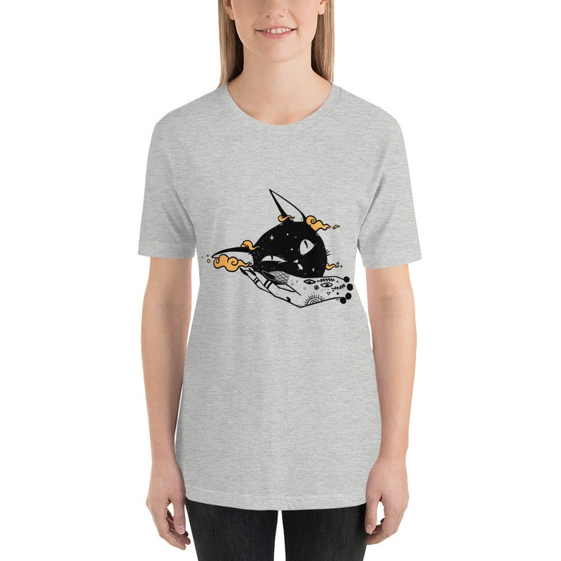 May include: Gray t-shirt with a black cat illustration. The cat is surrounded by orange clouds and is being held by a hand with a tattoo. The tattoo includes eyes, stars, and other symbols.