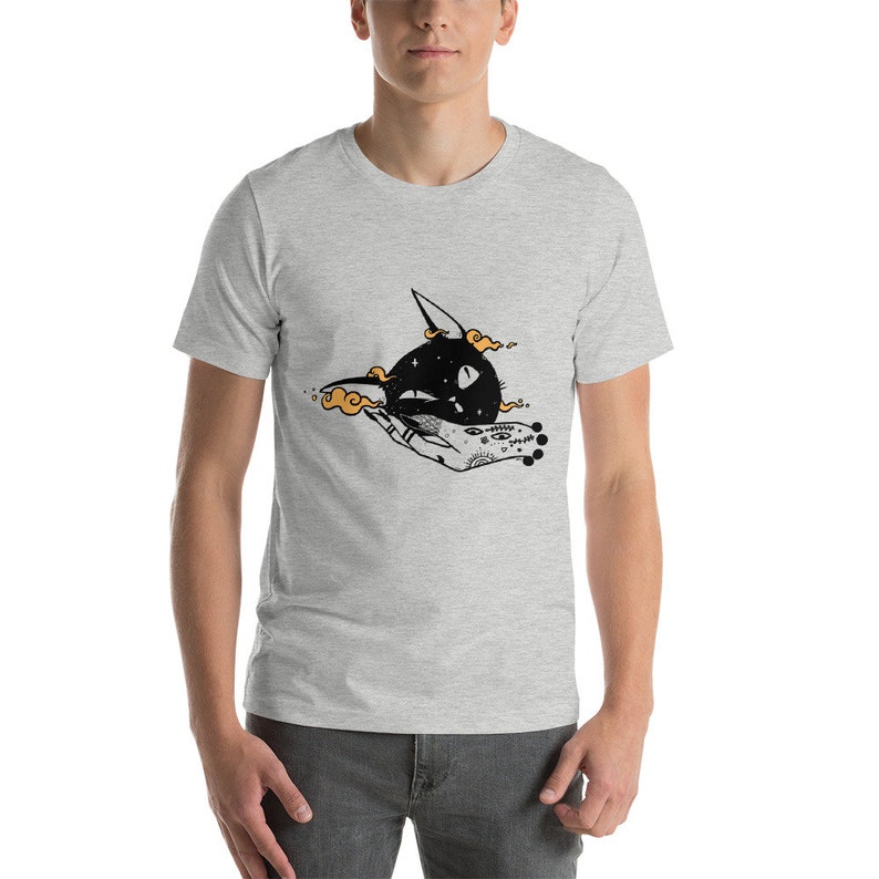May include: A gray t-shirt with a black and white illustration of a cat's head being held by a hand with a tattoo. The cat's eyes are white and the hand has a tattoo of a star, a circle, and a crescent moon.