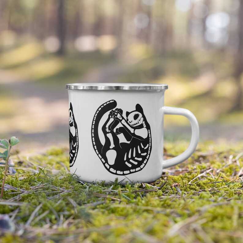 May include: White enamel mug with a black and white illustration of a cat skeleton curled up. The mug has a silver rim and handle.