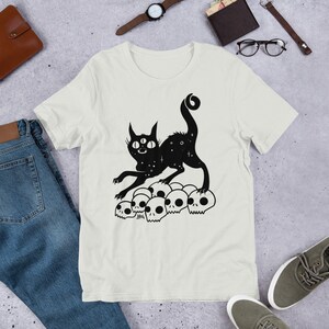 以下が含まれることがあります： 白いTシャツに、3つの目をした黒い猫のシルエットが、頭蓋骨の山の上に立っているデザインがプリントされています。