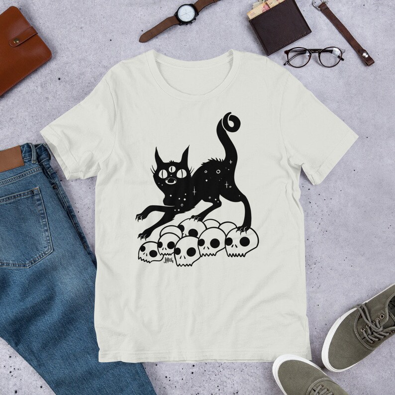 以下が含まれることがあります： 白いTシャツに、3つの目をした黒い猫のシルエットが、頭蓋骨の山の上に立っているデザインがプリントされています。