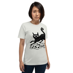 以下が含まれることがあります： 白いTシャツに、3つの目と星形の体を持つ黒い猫のシルエットが、頭蓋骨の山の上に立っているデザインがプリントされています。