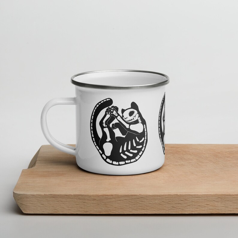 May include: White enamel mug with a black and white illustration of a cat skeleton. The cat is curled up in a ball with its tail wrapped around its body.