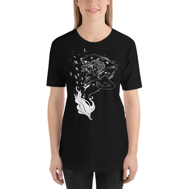 Op de afbeelding: Zwart T-shirt met een wit grafisch ontwerp van een vrouw met meerdere wolfskop en ogen. Het ontwerp is omringd door sterren en andere hemelse elementen.