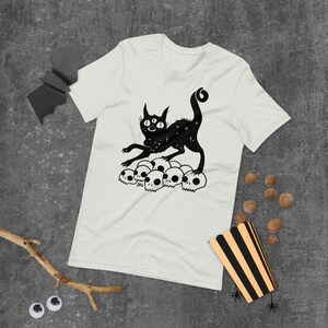 以下が含まれることがあります： 白いTシャツに、3つの目をした猫が頭蓋骨の山の上に立っている黒いイラストが描かれています。猫の体には星形の模様があります。