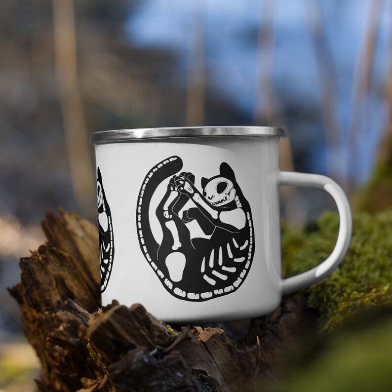 May include: White enamel mug with a black and white illustration of a cat skeleton. The cat is curled up in a circle with its tail wrapped around its body.