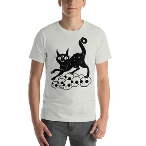 以下が含まれることがあります： 白いTシャツに、3つの目をした黒い猫のシルエットが、頭蓋骨の山の上に立っている。猫の体には星形の模様がある。