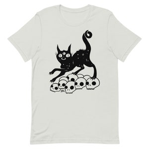以下が含まれることがあります： 3つの目と星形の体を持つ黒い猫が、白い頭蓋骨の山の上に立っています。猫は長い尻尾を持ち、上を見上げています。画像は白黒です。