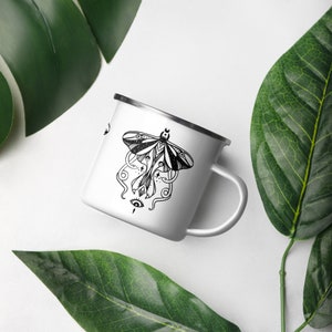 May include: White enamel mug with a black and white illustration of a moth with intricate details and a single eye. The moth has a crescent moon on its head.