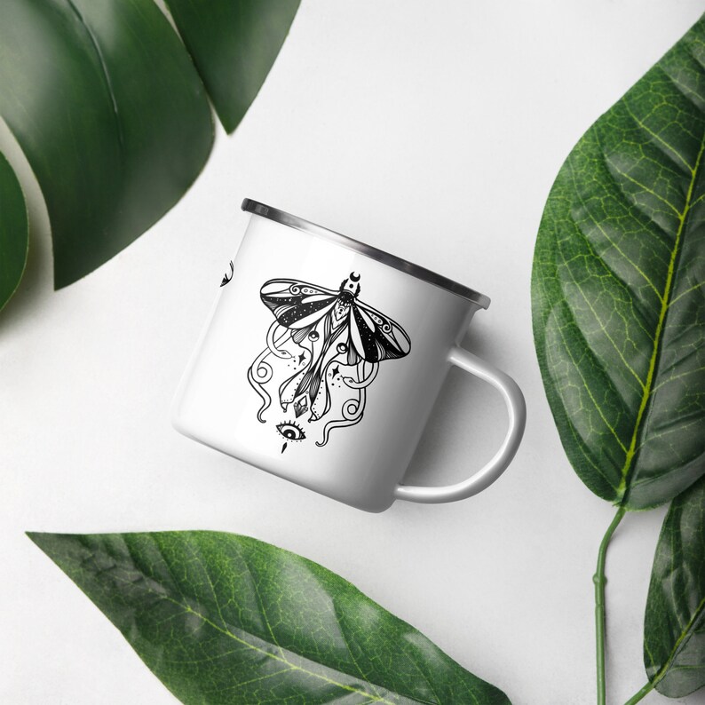 May include: White enamel mug with a black and white illustration of a moth with intricate details and a single eye. The moth has a crescent moon on its head.