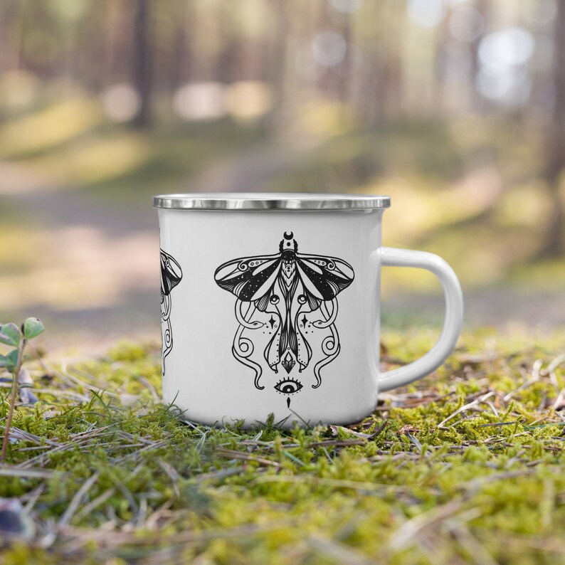 May include: White enamel camping mug with a black and white illustration of a moth with a third eye and a crescent moon on its head. The moth is surrounded by a swirling design.