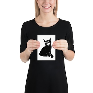 May include: Black and white art print featuring a whimsical illustration of a three-eyed cat with an open mouth. The cat is sitting upright, with a crescent moon shape above its head. The artwork is on a white background.