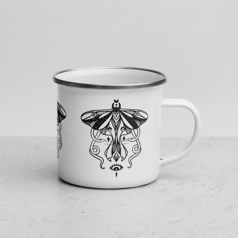May include: White enamel camping mug with a black and white illustration of a moth with a single eye in the center. The moth has intricate details and is surrounded by swirling lines.