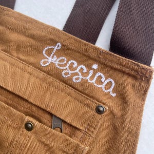 May include: Close-up of a brown canvas tote bag with a brown strap. The name "Jessica" is embroidered in white cursive on the bag. The bag has a zippered pocket and metal rivets.