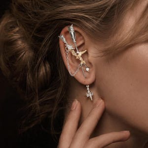 Könnte beinhalten: Ein silber- und goldfarbener Ohrring mit zwei pfeilförmigen Elementen, die durch eine feine Kette verbunden sind. Das Design umfasst eine goldfarbene Vogelfigur in der Mitte, kleine klare Steine und einen silberfarbenen Anhänger am Ohrläppchen.