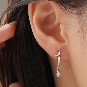Puede incluir: Un pendiente de tono plateado con forma de espada o daga en miniatura, que presenta un mango texturizado y una guarda. Una pequeña piedra preciosa transparente de talla marquesa cuelga de la punta de la hoja, creando un efecto colgante.