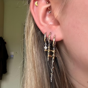 Può includere: Tre orecchini a cerchio color argento indossati sul lobo, ognuno con un ciondolo a forma di spada. I ciondoli presentano una finitura che mescola toni oro e argento. Un piccolo perno a mezzaluna dorata e un bilanciere curvo decorano l'orecchio.