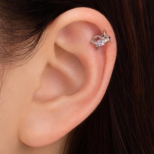 May include: A silver-toned ear cartilage stud featuring a star-shaped design with pointed rays and a central round, light purple gemstone. The jewelry is secured in the upper ear, showcasing a metallic finish and a delicate, symmetrical star motif.