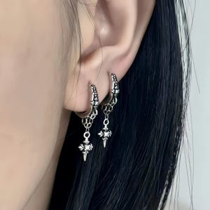 May include: A pair of silver-toned hoop earrings featuring a dangling cross-shaped charm with intricate filigree detailing and a small embedded clear stone. The hoops are decorated with a matching pattern and connected to the charms by thin chains.