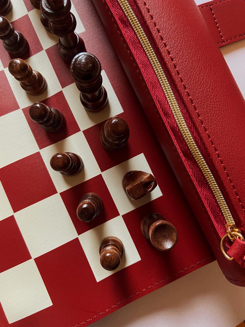May include: A red and white checkered chess board with wooden chess pieces. A red leather case with a gold zipper is visible. The chess pieces are dark brown and arranged on the board.