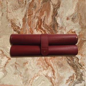May include: Two burgundy leather pen rolls, one atop the other, secured by a matching strap with a gold star accent. The rolls are cylindrical, with gold rivets at the ends. The background is a marble-like surface.