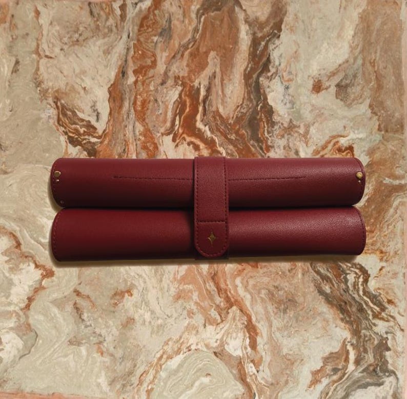 May include: Two burgundy leather pen rolls, one atop the other, secured by a matching strap with a gold star accent. The rolls are cylindrical, with gold rivets at the ends. The background is a marble-like surface.