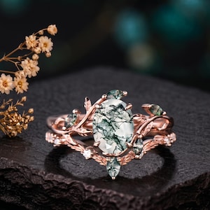 Könnte beinhalten: Ein Ring aus Roségold mit einem oval geschliffenen Moosachat-Mittelstein mit dunkelgrünen Einschlüssen. Die Ringschiene ist mit detaillierten Blatt- und Rankenmotiven gestaltet, die durch kleinere marquise-geschliffene Moosachatsteine ergänzt werden.