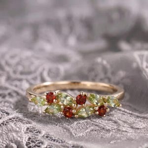 Könnte beinhalten: Ein goldener Ring mit einer Anordnung aus hellgrünen Edelsteinen im Marquise-Schliff und tiefroten Edelsteinen im Rundschliff, die in einem blattartigen Muster auf der Oberseite des Rings angeordnet sind und einen lebendigen Kontrast bilden.