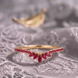 Könnte beinhalten: Ein goldener V-förmiger Ring mit einer Reihe kleiner runder roter Steine entlang der Ringschiene und fünf im Marquise-Schliff geschliffenen roten Steinen, die in der Mitte fächerförmig angeordnet sind. Der Ring hat ein poliertes Gold-Finish.