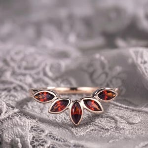 May include: A rose gold ring featuring five marquise-cut deep red garnet gemstones arranged in a curved, fan-like cluster. The band is thin and polished, showcasing a symmetrical design where the central stone is the largest, flanked by four smaller stones.