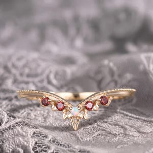 Könnte beinhalten: Ein goldener V-förmiger Ring mit einem zentralen runden Opal, flankiert von vier kleinen runden roten Granat-Edelsteinen. Das Band hat ein strukturiertes, spitzes Design, das der Chevron-Form folgt, wobei die Steine entlang der Vorderkante sitzen.