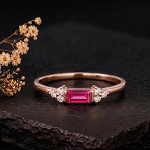 Könnte beinhalten: Ein Ring aus Roségold mit einem zentralen, im Baguetteschliff geschliffenen rosa Edelstein, der auf jeder Seite von drei kleinen, runden, klaren Steinen flankiert wird. Das Band ist schmal und poliert und betont den leuchtend rosa Stein.