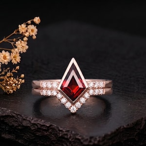Könnte beinhalten: Ein Ring-Set aus Roségold mit einem tiefroten, drachenförmigen Granat als Mittelstein in einer Zargenfassung. Sowohl die Ringschiene als auch der passende chevronförmige Vorsteckring sind mit kleinen, runden, klaren weißen Steinen besetzt.
