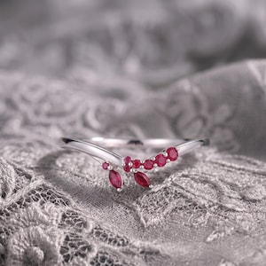Könnte beinhalten: Ein V-förmiger Ring in Silberoptik mit einer Ansammlung kleiner, runder und marquise-geschliffener roter Rubine, die in einem zarten, asymmetrischen Muster entlang der Ringschiene angeordnet sind und ein poliertes Metallfinish aufweisen.