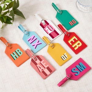 May include: A collection of colorful luggage tags with various initials. The tags are in shades of orange, pink, yellow, red, green, and blue. Each tag has a matching strap and buckle. The initials are in a contrasting color.