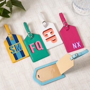 Op de afbeelding: Een verzameling gepersonaliseerde bagagelabels in geel, groen, wit en roze. Elke tag heeft unieke initialen. Eén tag is open en toont een blanco inzetstuk. De tags zijn gerangschikt op een wit oppervlak.
