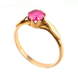 May include: A gold ring featuring a round, faceted, vibrant pink gemstone set in a six-prong crown setting. The band is smooth and polished, with tapered shoulders that lead up to the central stone, creating a clean and classic jewellery design.