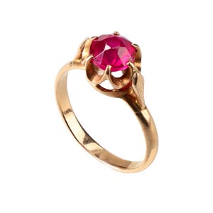 May include: A gold ring featuring a round, faceted, vibrant pinkish-red gemstone set in a six-prong crown-style mounting. The band is smooth and polished, connecting to the setting with a decorative, petal-like metal structure surrounding the stone.