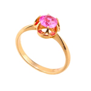 Può includere: Un anello in oro con una pietra preziosa rotonda, sfaccettata e di colore rosa brillante, incastonata in una montatura a sei griffe a forma di corona. Il gambo è liscio e lucido, con un design semplice che si restringe verso la pietra.
