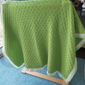 Puede incluir: Una manta de punto verde brillante con un patrón cuadrado texturizado. La manta tiene un ribete de encaje blanco y verde claro a lo largo de los bordes. La manta está sobre un marco de madera.